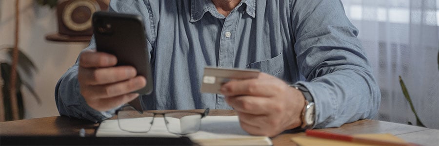 Article teaser image. Photo of holding a mobile device and credit card with financial documents spread out on a kitchen table.