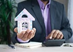 Man in suit holding a model house in one hand while typing in a calculator in the other hand